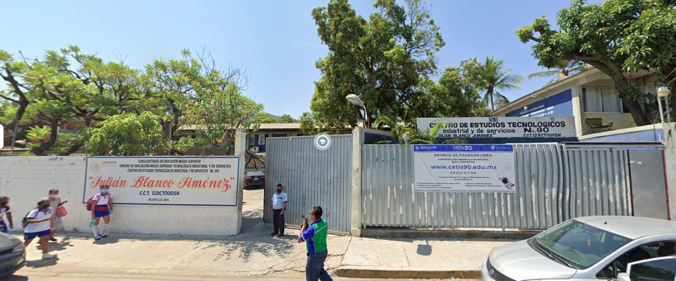 Fotografía en la que se muestra la entrada del plantel, conformada por un muro blanco en el que está escrito el nombre del plantel. Del lado derecho del muro hay dos rejas y la que está enseguida del muro es la que permite el acceso a las instalaciones.